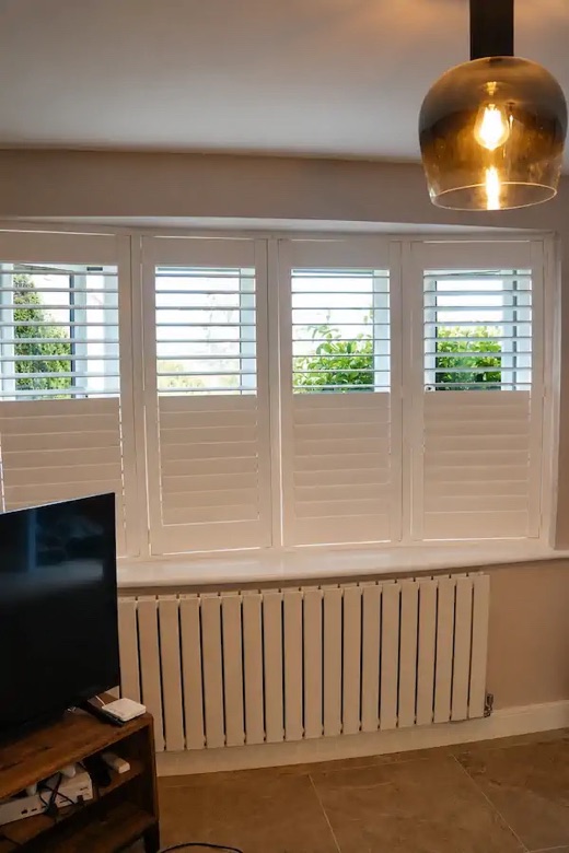 Living-room four-panel bay window in Stourbridge with full height white shutters, louvres tilted open showing the garden beyond