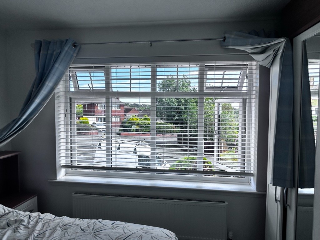 Snow White wood Venetian blinds on a bedroom window with blue check curtains, slats open