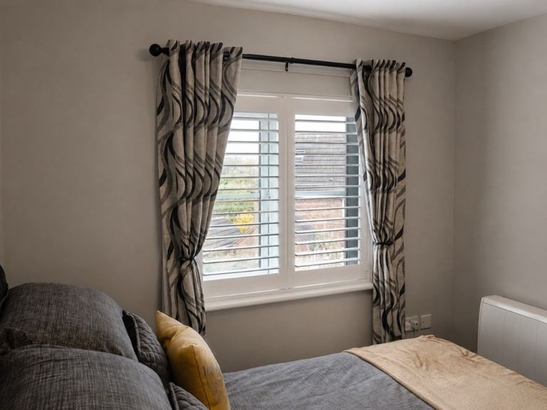 Full height plantation shutters fitted to a bedroom window in Tipton, louvres tilted open with patterned curtains tied back either side
