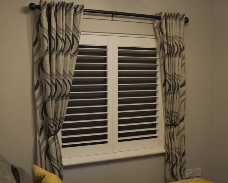 Shutter & Shade in a West Midlands bedroom: white plantation shutters closed, integrated blackout blind pulled down behind, visible as a dark layer through the open louvres, curtains drawn back either side
