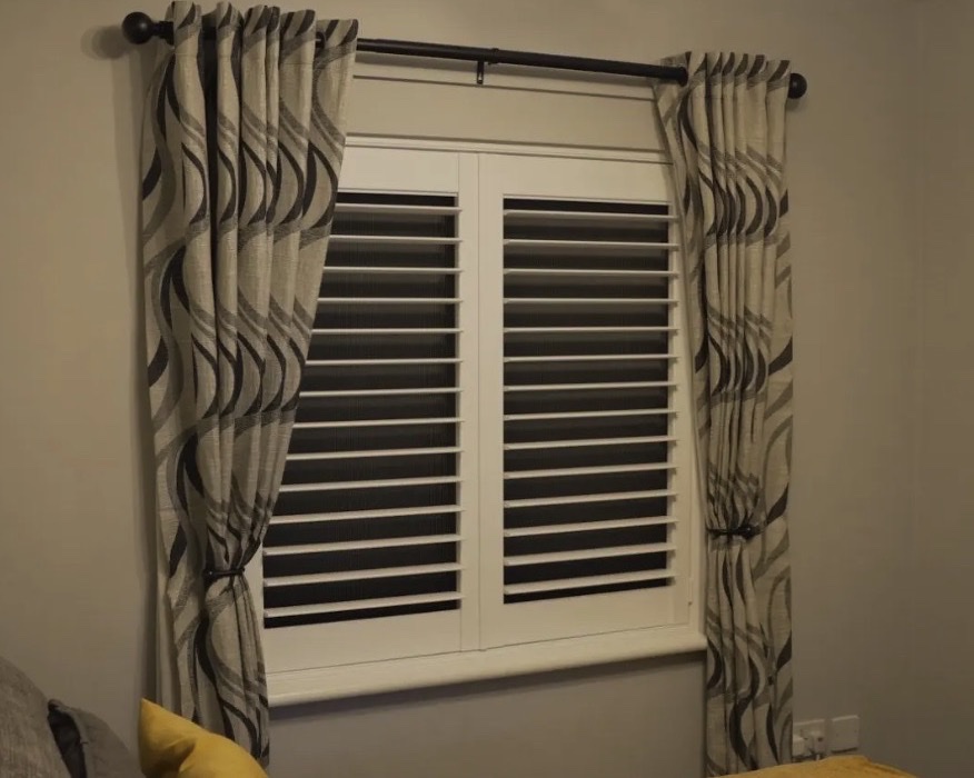 Shutter & Shade in a West Midlands bedroom: white plantation shutters closed, integrated blackout blind pulled down behind, visible as a dark layer through the open louvres, curtains drawn back either side
