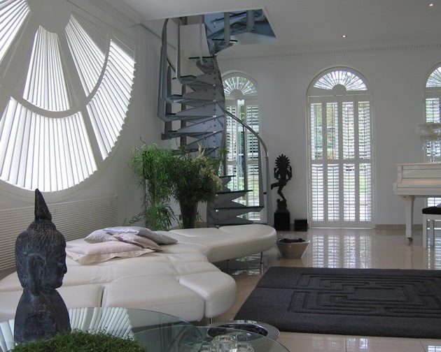 Open-plan West Midlands home with three different special shapes by Greystone Shutters: a fan-pattern circular shutter on a porthole window, plus tall arched-top shutters across the rear elevation, louvres set to filter daylight