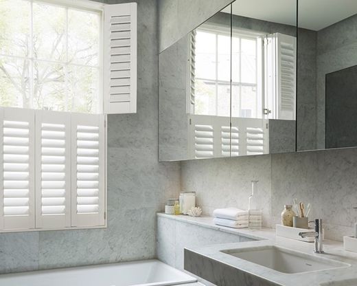 Tier-on-tier plantation shutters in a marble bathroom, top tier folded open against the reveal for a treetop view while the bottom tier louvres stay closed for bath-time privacy