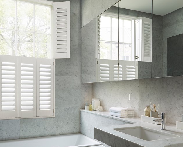Tier-on-tier plantation shutters in a marble bathroom, top tier folded open against the reveal for a treetop view while the bottom tier louvres stay closed for bath-time privacy